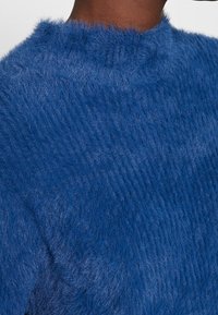 Pull bleu duveteux à col montant, avec une texture douce et une coupe légèrement oversize, mettant en valeur une couleur profonde et riche.