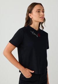 Vrouw in een zwart kortärmet-shirt met een klein rood roosje en een wit steel ontwerp nabij de halslijn, hand in de zak.