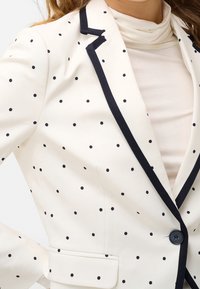 Bílý blazer s černými puntíky, námořnickými detaily, zapínáním na jeden knoflík a bočními kapsami. Hladká tkanina se strukturovaným střihem.