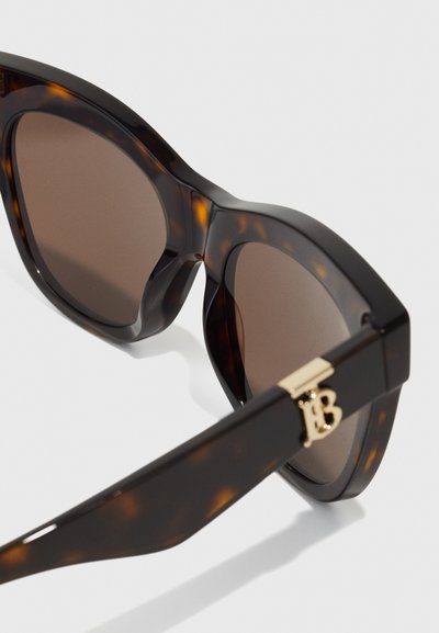 Lunettes de soleil écaille de tortue marron avec des verres foncés, large monture et ferrures dorées comportant un détail de logo sur la branche.