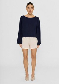 Marineblauwe geribde long-sleeve top, gecombineerd met beige shorts. De top heeft een wijde halslijn en gestructureerde mouwen; de schoenen zijn licht van kleur en hebben uitsparingen.