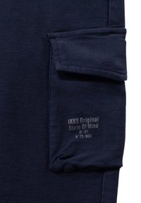 Donkerblauwe katoenstof met een subtiele textuur. Bevat een zak en een geperst logo-tekst, "IKKS Original State Of Mind," op het materiaal.