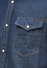 Tamsiai mėlyna džinsinio audinio marškiniai su kišene ir baltais spaudėmis. Yra oranžinė siūlė ir etiketė ant kišenės.