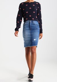 Jeanspencilkjol, blekt blå med fransad fåll, i kombination med en svart långärmad topp med röda blommönster och bärs med svarta sneakers.