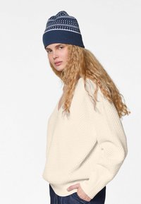 Cremfarbener, strukturierter Strickpullover mit V-Ausschnitt und lockerer Passform, kombiniert mit einer marineblauen Mütze mit weißen Musterakzenten.