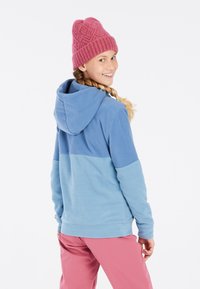Un hoodie en polaire bleu clair avec une partie supérieure bleu foncé, doté d'une capuche avec cordon de serrage. Associé à un pantalon rose et un bonnet rose texturé.