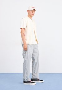 Light beige sleeveless vest over a white t-shirt, gray cargo pants with side pockets, and black sneakers with pink laces.