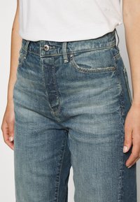 Kädet lanteilla, päällä haalistuneet siniset farkut, joissa näkyvät napit ja taskut, yhdistettyinä valkoiseen sisään laitettuun paitaan.