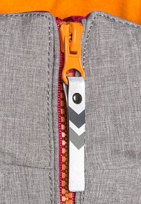 Szary materiał z wyraźnym pomarańczowym uchwytem suwaka. Taśma suwaka ma kontrastowe różowe wykończenie, a srebrna metka ma wzór w zygzaku w czerni i bieli.