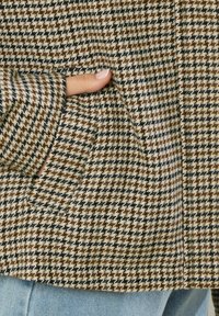 Giacca con motivo houndstooth in tessuto marrone, nero e crema, dotata di tasca laterale e maniche con risvolto; vista ravvicinata che mette in evidenza i dettagli del tessuto.