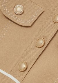 Bež kudumaterjal, millel on nähtavad õmblused, nelja kuldse tooni nupuga, millel on tekstureeritud äär ja valge riba ääres.