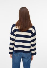Personne aux longs cheveux bruns portant un pull à rayures horizontales bleu marine et blanc et un jean bleu, debout, de dos sur un fond uni.