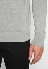 Gray knit sweater with ribbed cuffs and hem, featuring a soft texture. Worn with black pants, showing the lower arm and hand.