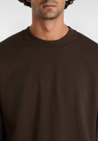 Mies, joka on pukeutunut tummanruskeaan pyöreäkauluksiseen collegepaitaan, paljastaen kaulan, leukalinjan ja osittain huulia yksinkertaista taustaa vasten.