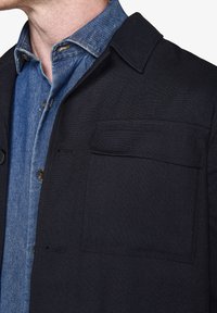 Sort stofjakke med en struktureret krave, der har en brystlomme og er parret med en blå denim skjorte med knapper under.