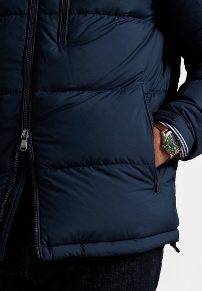 Marineblå puffjakke med kviltedesign, frontlynlås, sidelommer og elastisk kant, parret med et grønt ur på håndleddet.
