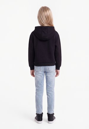 Enfant aux longs cheveux blonds portant un sweat à capuche noir, un jean bleu clair et des baskets montantes noires, debout dos à un fond blanc.