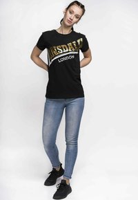 Black t-shirt featuring gold and white lettering "Lonsdale London." Paired with fitted blue jeans and black athletic shoes.