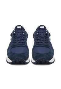 Tumši zili sporta apavi ar zamšādas materiālu, teksturētiem akcentiem, baltiem elementiem un gumijas zolīti ar viļņotu rakstu.