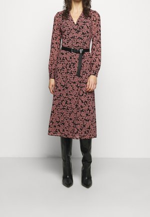 Kvinde iført en sort midi-kjole med lyserøde blomsterprint, lange ærmer, sort bælte og knæhøje sorte støvler, stående op ad en hvid væg.
