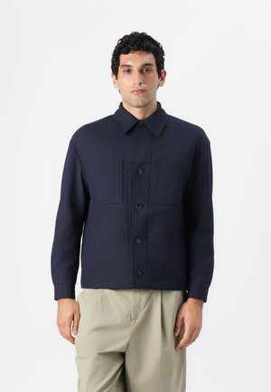 Navy blue jacket, made of thick fabric, featuring a collar, button closure, and two front pockets. Worn with light beige trousers.