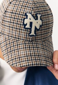 Braune und beige Kappe mit Hahnentrittmuster und einem navyblauen, bestickten Logo. Verfügt über eine gebogene Krempe und eine strukturierte Stoffdetail.