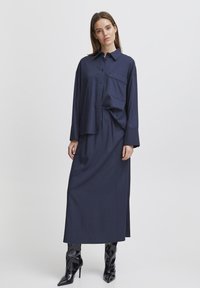Chemise bleue marine à manches longues et à boutons assortie à une longue jupe. Tissu lisse, coupe décontractée, avec une poche poitrine. Bottines à talons noires.