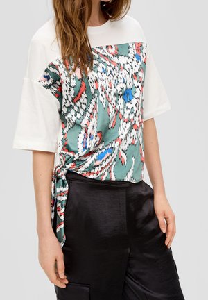 Kobieta ubrana w biały top z krótkim rękawem z abstrakcyjnym nadrukiem w kolorach zielonym, czerwonym, niebieskim i białym oraz czarne satynowe spodnie z wysokim stanem.