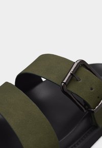 Κοντινό πλάνο μαύρης σαγιονάρας με δύο φαρδιές χρυσόπρασινες λωρίδες, η μία με μαύρη μεταλλική αγκράφα για ρύθμιση.