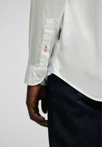 Witte langemouwen shirt met een textuurafwerking, voorzien van blauwe en rode knopen op de manchetten en een zijsplit, gecombineerd met donkere blauwe jeans.