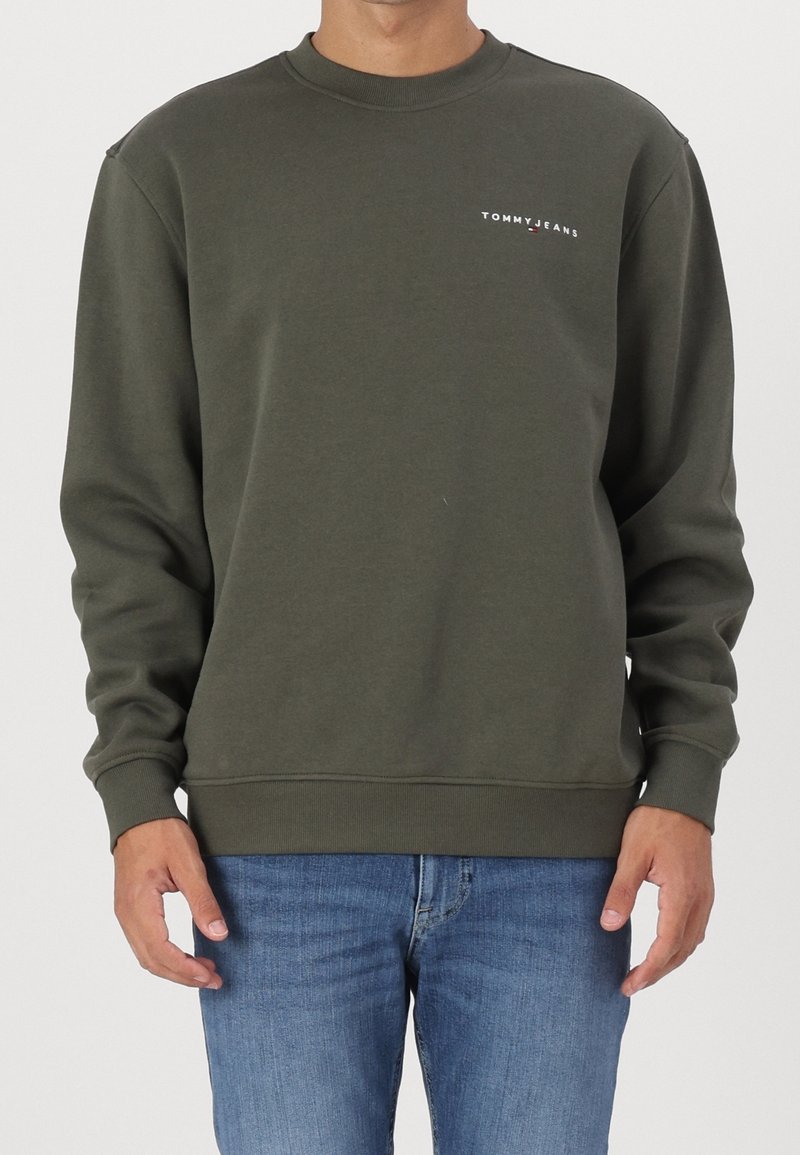 Man wearing olive green Tommy Jeans sweatshirt and blue jeans, standing against a plain background.