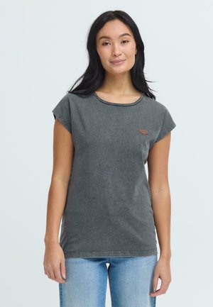 Femme aux longs cheveux noirs portant un haut sans manches gris foncé et un jean bleu clair, debout devant un fond clair uni.