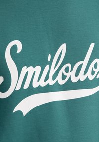 Camiseta de algodón en color azul verdoso con un gran texto en escritura retro blanca que dice "Smilodex" y una línea curvada debajo, mostrando una textura suave y mate.