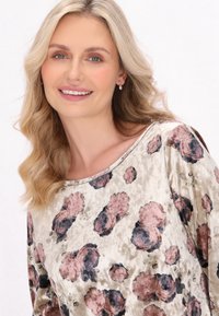 Hosszú ujjú, környakú felső puha arany szövetből, rózsás mintával, amelyen rózsaszín és sötétkék rózsák találhatók, kiemelve a texturált felületet.