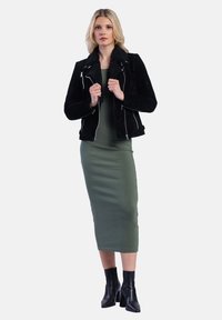 Žena v černé bundě s beránkovým límcem, sebevědomě stojící v zelených šatech a černých kotníkových botách na jednotném pozadí.