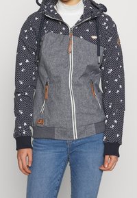 Veste à capuche avec tissu bleu marine à pois et motif floral sur les manches, corps gris, fermeture éclair et accents marron sur les poches.