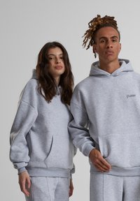 Pilki gobtuvai, pagaminti iš minkštos medžiagos, su kengūros kišene, gaubtais rankogaliais ir apačia. Vienas turi spausdintą logotipą ant krūtinės.