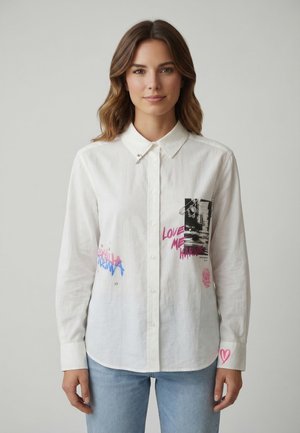 Donna con capelli castani alla lunghezza delle spalle indossa una camicia bianca con bottoni e una scritta grafica colorata, e jeans azzurri chiari, in piedi davanti a uno sfondo semplice.