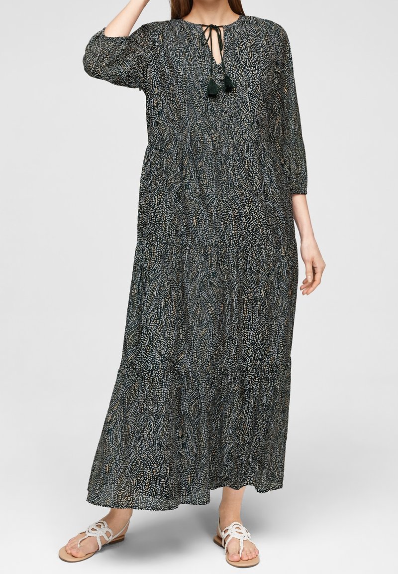 Femme portant une longue robe noire et blanche à motifs avec des manches 3/4 et des sandales plates blanches tressées sur un fond uni.