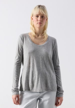 Blond naine, kellel on seljas helehall pika varrukaga särk ja helesinised teksad, seisab ühevärvilise valge tausta ees.