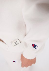 Beige sweatshirt och träningsbyxor med Champion-logotyper på ärmen och benet, samt en patch med texten "Var din egen mästare" nära midjan.