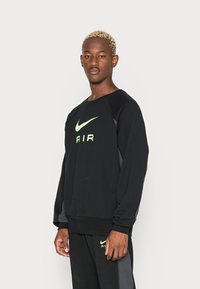 Melnā sporta krekls ar apaļu kakla izgriezumu un garām piedurknēm, kuram ir fluorescējoši zaļa Nike logo un "AIR" teksts, izgatavots no mīksta auduma.