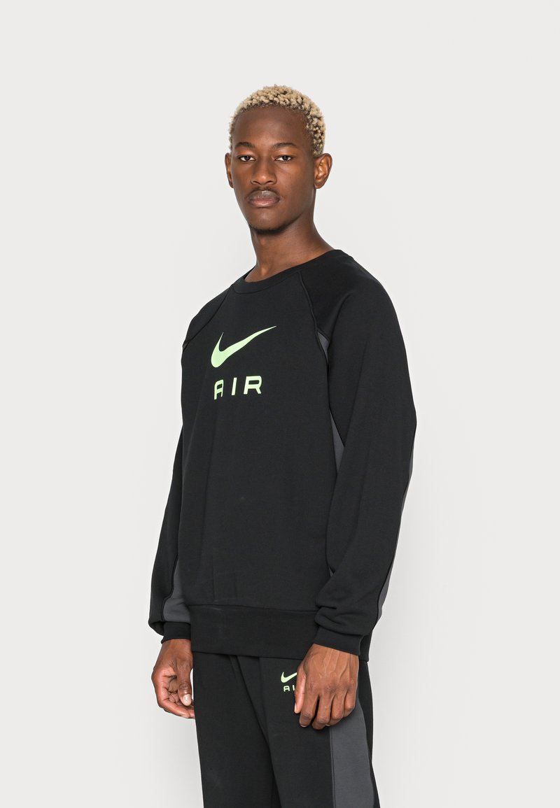 Melnā sporta krekls ar apaļu kakla izgriezumu un garām piedurknēm, kuram ir fluorescējoši zaļa Nike logo un "AIR" teksts, izgatavots no mīksta auduma.