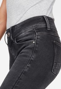 Vaqueros negros de denim de tiro alto con una textura suave, un leve desgaste y un diseño clásico de cinco bolsillos; detalles de botón y remaches metálicos.