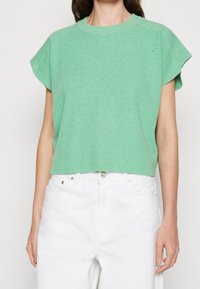 Zielony prążkowany krótki rękawowy sweter z okrągłym dekoltem oraz lekko rozszerzanymi rękawami, zestawiony z białymi spodniami o wysokim stanie.