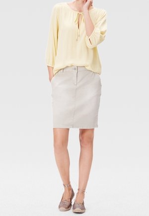 Kvinde iført en lys gul bluse, beige knælang nederdel og brune espadrillosko med ankelbånd, stående med den ene hånd i lommen.