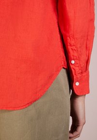 Red linen shirt with a curved hem, long sleeves, and buttoned cuffs, paired with light olive green pants. Smooth texture, casual fit.