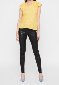 Blusa floreale gialla con maniche corte a volant, nodo al collo; abbinata a pantaloni di pelle nera attillati e scarpe con tacco azzurro chiaro.