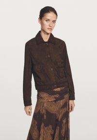 Bruine suède jas met een kraag, voorzakken en knopen. Draagt over een bruin patroonrok met gouden accenten.