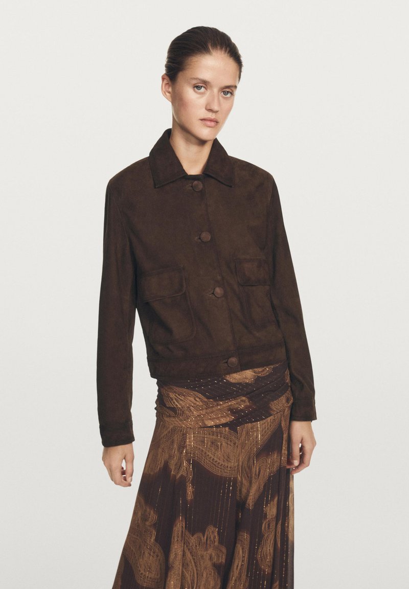 Bruine suède jas met een kraag, voorzakken en knopen. Draagt over een bruin patroonrok met gouden accenten.